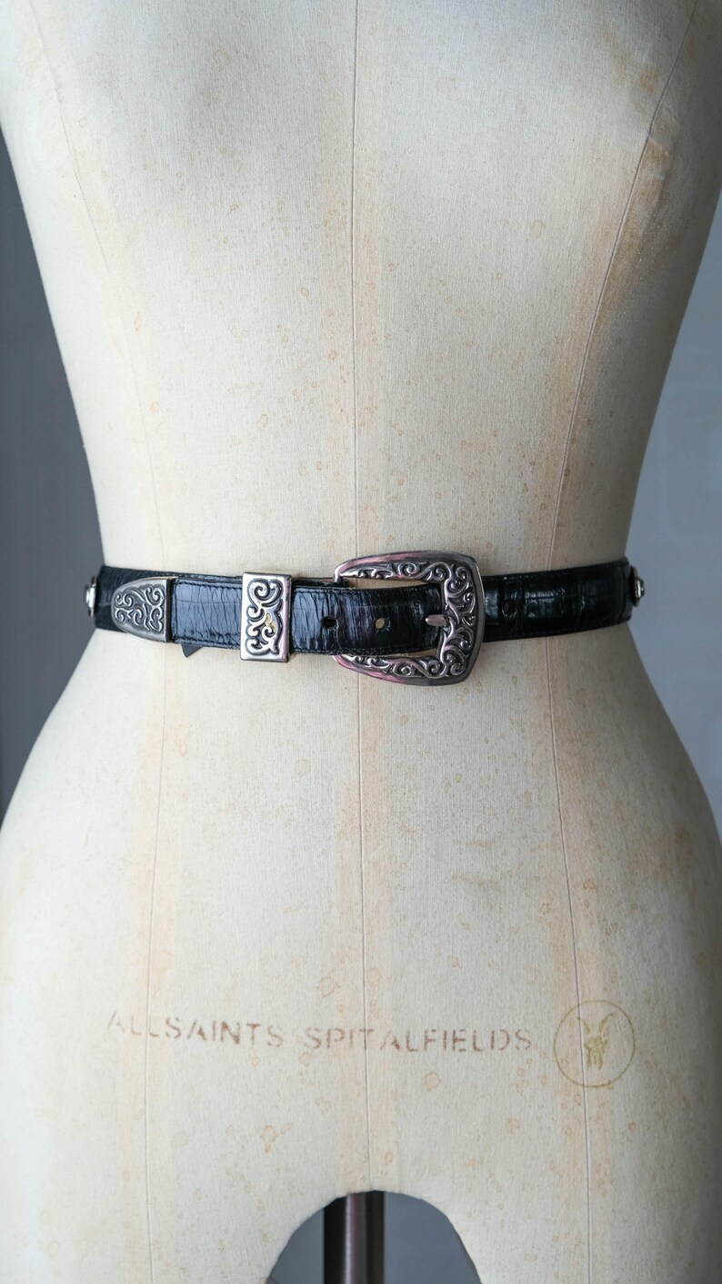 May include: Black belt with a silver buckle and accents. The belt is made of a textured material and features a decorative buckle with an ornate design. The belt is displayed on a mannequin.