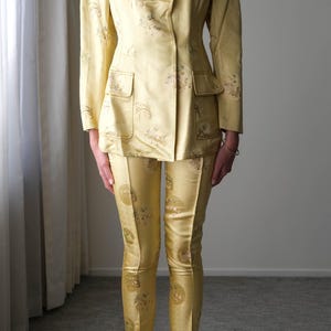 May include: A vintage yellow suit featuring a double-breasted blazer with two front pockets and matching tailored pants. The suit has a subtle floral pattern and is made of a shiny fabric. The pants are fitted and the suit is a classic style.