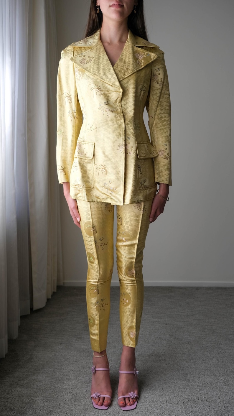 May include: A vintage yellow suit featuring a double-breasted blazer with two front pockets and matching tailored pants. The suit has a subtle floral pattern and is made of a shiny fabric. The pants are fitted and the suit is a classic style.