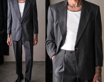 May include: A charcoal gray suit with a single-button blazer and matching trousers. The suit is paired with a white undershirt and black boots. The suit has a classic, tailored fit, suitable for formal occasions.