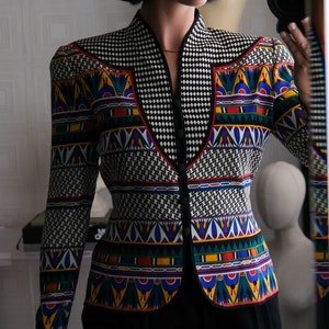May include: A woman wearing a black and white patterned blazer with colorful geometric designs. The blazer has a high collar and a button closure. The woman is wearing black pants.