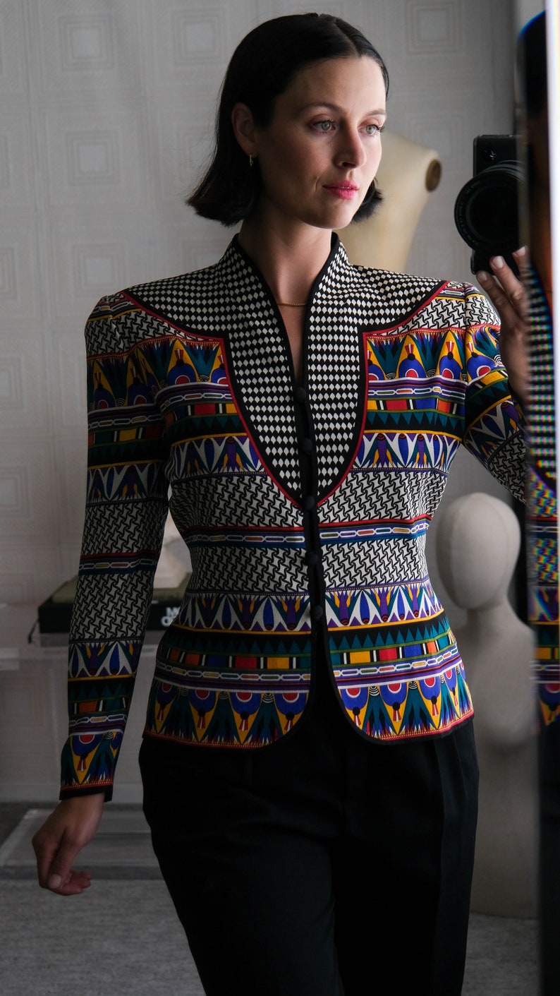 May include: A woman wearing a black and white patterned blazer with colorful geometric designs. The blazer has a high collar and a button closure. The woman is wearing black pants.