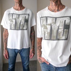 May include: A white short-sleeve t-shirt featuring a large graphic print on the chest. The graphic consists of the letters NYC in a bold, block font, with black and white photographic imagery of New York City landmarks integrated within the letters.