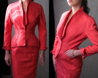 May include: A coral-colored two-piece suit featuring a fitted blazer with a unique collar and three buttons, paired with a matching knee-length skirt. The suit has long sleeves with buttoned cuffs. The fabric appears to have a subtle texture.