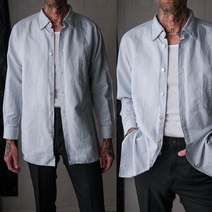 May include: A light blue button-down shirt with long sleeves and a chest pocket. The shirt is worn over a white undershirt and paired with black pants. The shirt has a classic collar and buttoned cuffs.