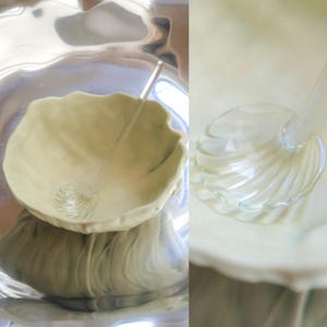 Könnte beinhalten: Eine blassgrüne Keramikschale mit gewelltem, blattartigem Rand, kombiniert mit einer durchsichtigen Glasschöpfkelle mit muschelförmigem Laffe. Die Kelle hat einen langen, schlanken Stiel und liegt in der Schale, was ihr transparentes Design zeigt.