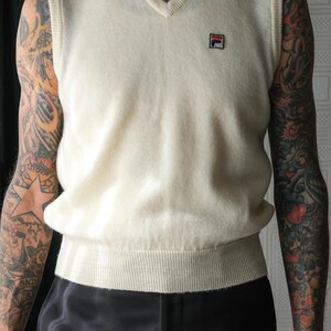 Peut inclure: Gilet sans manches cr&egrave;me &agrave; col en V avec un ourlet c&ocirc;tel&eacute; et un petit &eacute;cusson logo. Le gilet est sans manches et a une coupe d&eacute;contract&eacute;e. Le mod&egrave;le porte un pantalon noir.