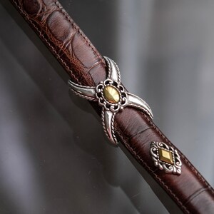 K&ouml;nnte beinhalten: Ein brauner Lederg&uuml;rtel mit Krokodiltextur. Der G&uuml;rtel hat eine silber- und goldfarbene Schnalle mit dekorativem Design. Ein klassisches Accessoire f&uuml;r jede Garderobe.