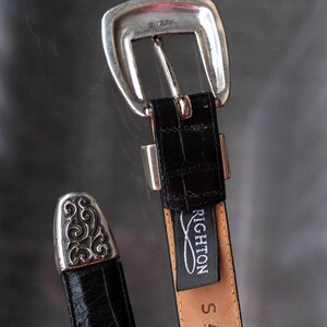 May include: A black leather belt with a silver buckle and tip. The belt has a crocodile texture and a "Brighton" tag. The belt is size S and has the number 44403 printed on it. The buckle is silver-toned.