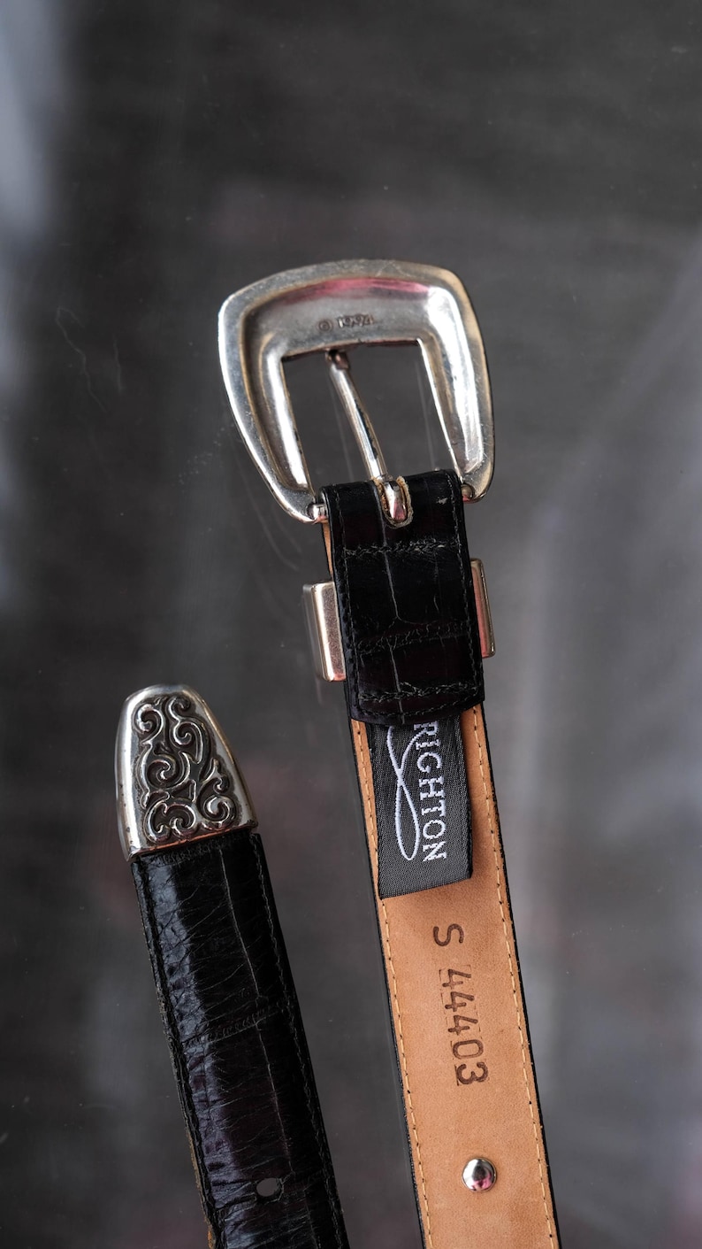 May include: A black leather belt with a silver buckle and tip. The belt has a crocodile texture and a "Brighton" tag. The belt is size S and has the number 44403 printed on it. The buckle is silver-toned.
