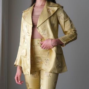 May include: A golden yellow suit featuring a blazer with two front pockets and matching pants. The suit has a floral pattern and a textured appearance. The blazer has a notched collar and the pants have a button closure.