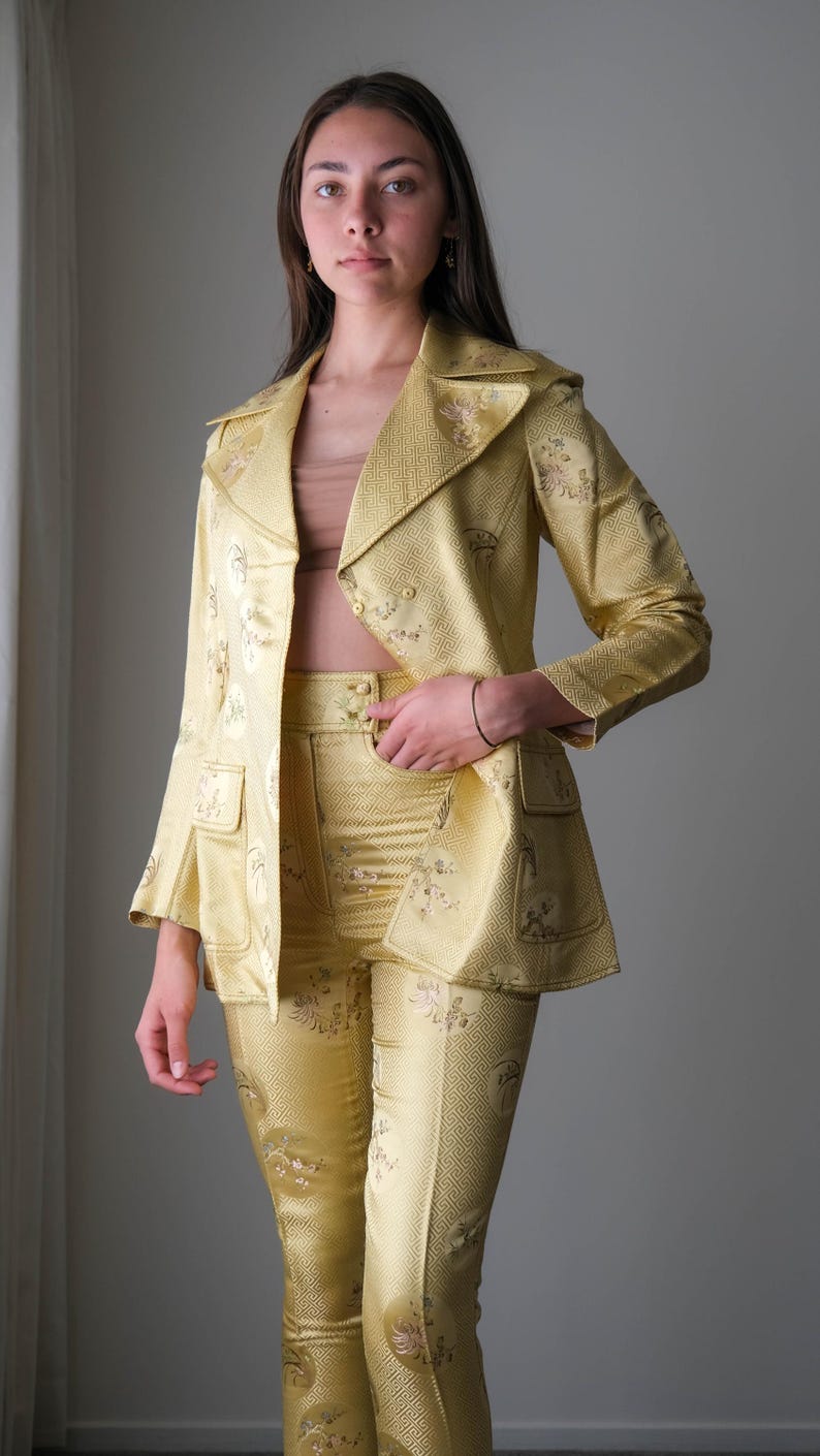 May include: A golden yellow suit featuring a blazer with two front pockets and matching pants. The suit has a floral pattern and a textured appearance. The blazer has a notched collar and the pants have a button closure.