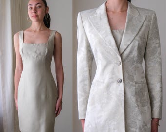 May include: A two-piece ensemble featuring a light beige sheath dress with a square neckline and a matching floral-patterned blazer. The blazer has a single-button closure and a notched lapel. The dress and blazer are made of a textured fabric.