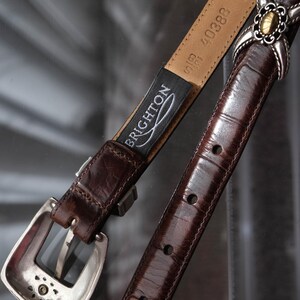 K&ouml;nnte beinhalten: Ein brauner Lederg&uuml;rtel mit silberner Schnalle und dekorativen Akzenten. Der G&uuml;rtel hat eine strukturierte Oberfl&auml;che und ein Etikett mit der Aufschrift "BRIGHTON". Der G&uuml;rtel ist etwa 2,5 cm breit.