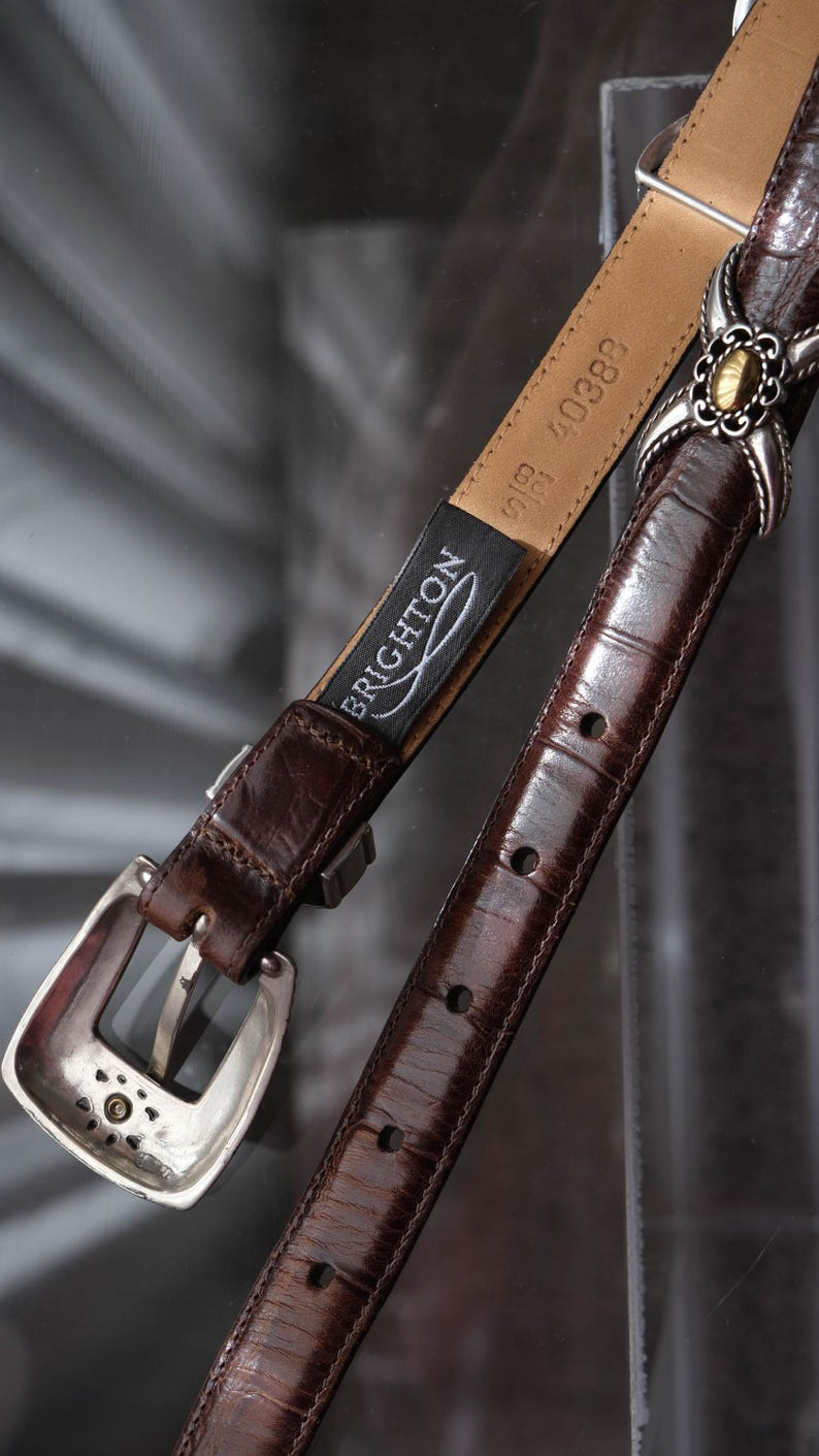 K&ouml;nnte beinhalten: Ein brauner Lederg&uuml;rtel mit silberner Schnalle und dekorativen Akzenten. Der G&uuml;rtel hat eine strukturierte Oberfl&auml;che und ein Etikett mit der Aufschrift "BRIGHTON". Der G&uuml;rtel ist etwa 2,5 cm breit.