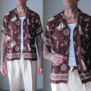 Puede incluir: Una camisa de manga corta con botones en color marrón oscuro que presenta un estampado náutico repetitivo de veleros, faros, timones y anclas en blanco. La prenda incluye un cuello de muesca, un bolsillo en el pecho y un borde con diseño de cuerda.