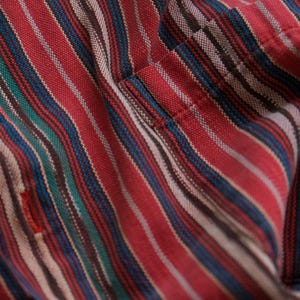 May include: Close-up of a red striped fabric with blue, green, and white stripes. The fabric has a textured appearance and a small red detail. The stripes run diagonally across the frame, creating a dynamic pattern. The fabric appears to be part of a shirt.