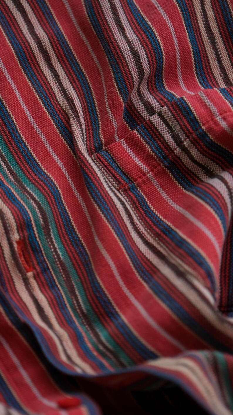 May include: Close-up of a red striped fabric with blue, green, and white stripes. The fabric has a textured appearance and a small red detail. The stripes run diagonally across the frame, creating a dynamic pattern. The fabric appears to be part of a shirt.