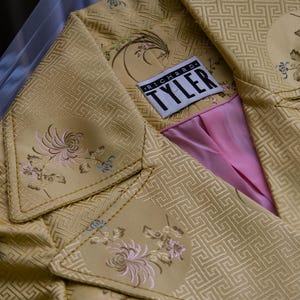 May include: A close-up of a gold-colored jacket with a geometric pattern and floral embroidery. The jacket has a pink lining and a label that reads "RICHARD TYLER". The jacket is displayed on a clear surface.