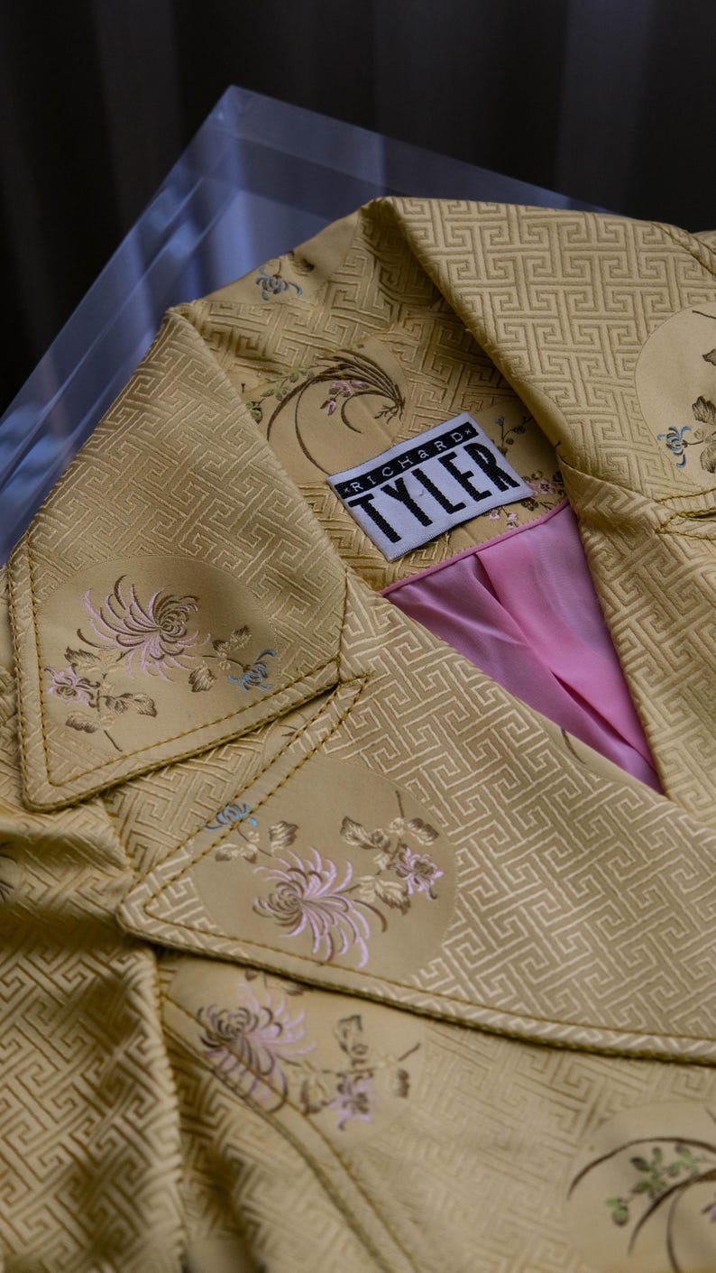 May include: A close-up of a gold-colored jacket with a geometric pattern and floral embroidery. The jacket has a pink lining and a label that reads "RICHARD TYLER". The jacket is displayed on a clear surface.
