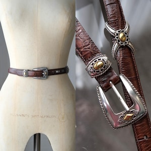 K&ouml;nnte beinhalten: Ein brauner Lederg&uuml;rtel mit silberner Schnalle und dekorativen Akzenten. Der G&uuml;rtel hat ein strukturiertes Muster und goldfarbene Details. Die Schnalle ist silberfarben mit einem goldfarbenen Mittelteil. Der G&uuml;rtel wird auf einer Schaufensterpuppe pr&auml;sentiert.