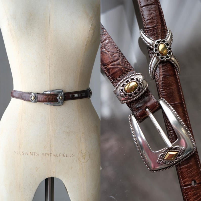 K&ouml;nnte beinhalten: Ein brauner Lederg&uuml;rtel mit silberner Schnalle und dekorativen Akzenten. Der G&uuml;rtel hat ein strukturiertes Muster und goldfarbene Details. Die Schnalle ist silberfarben mit einem goldfarbenen Mittelteil. Der G&uuml;rtel wird auf einer Schaufensterpuppe pr&auml;sentiert.