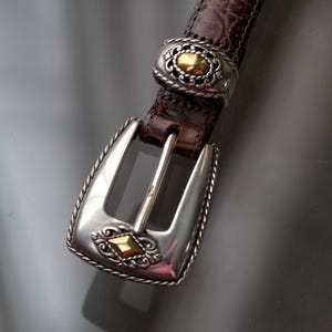 K&ouml;nnte beinhalten: Brauner Lederg&uuml;rtel mit einer strukturierten, krokodilartigen Pr&auml;gung. Der G&uuml;rtel hat eine silberfarbene Schnalle und goldfarbene Akzente. Die Schnalle hat ein verziertes Design.