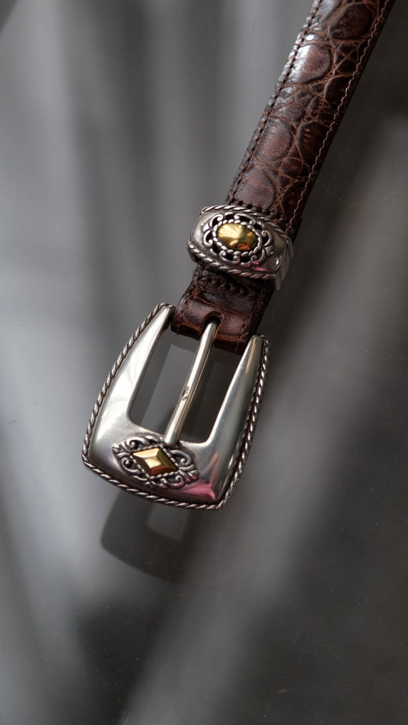 K&ouml;nnte beinhalten: Brauner Lederg&uuml;rtel mit einer strukturierten, krokodilartigen Pr&auml;gung. Der G&uuml;rtel hat eine silberfarbene Schnalle und goldfarbene Akzente. Die Schnalle hat ein verziertes Design.