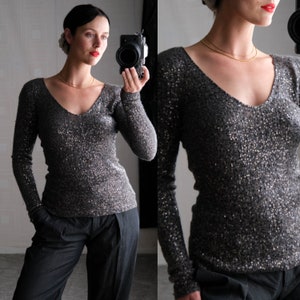 May include: A long-sleeved, dark gray sequined top with a V-neck. The top is covered in small, reflective sequins, creating a shimmering effect. The sleeves are long and fitted, and the overall style is elegant and suitable for evening wear.