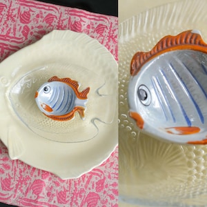 May include: A clear glass fish-shaped plate with a textured scale pattern, featuring a smaller ceramic fish-shaped bowl painted in light blue, white, and orange. The ceramic bowl has a detailed eye, fins, and a distinct orange-colored dorsal fin.