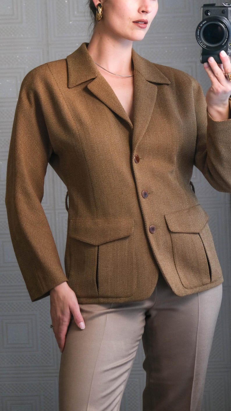 May include: A brown blazer with a classic design, featuring a notched collar, two front pockets, and button closures. The blazer is paired with tan-colored trousers. The fabric has a subtle textured pattern.