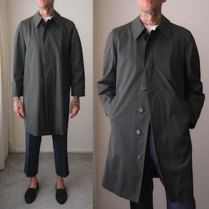 May include: A dark olive green trench coat featuring a classic point collar, long sleeves, and a front button closure. The coat has a smooth, solid finish and a straight-cut silhouette that falls to the mid-thigh, providing a clean and structured look.