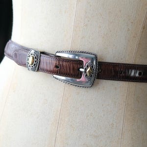 K&ouml;nnte beinhalten: Ein brauner Lederg&uuml;rtel mit silberner Schnalle und Akzenten. Der G&uuml;rtel hat ein strukturiertes, krokodilartiges Muster. Die Schnalle hat einen silbernen Rahmen mit goldfarbenen Details. Der G&uuml;rtel wird auf einer Schaufensterpuppe pr&auml;sentiert.