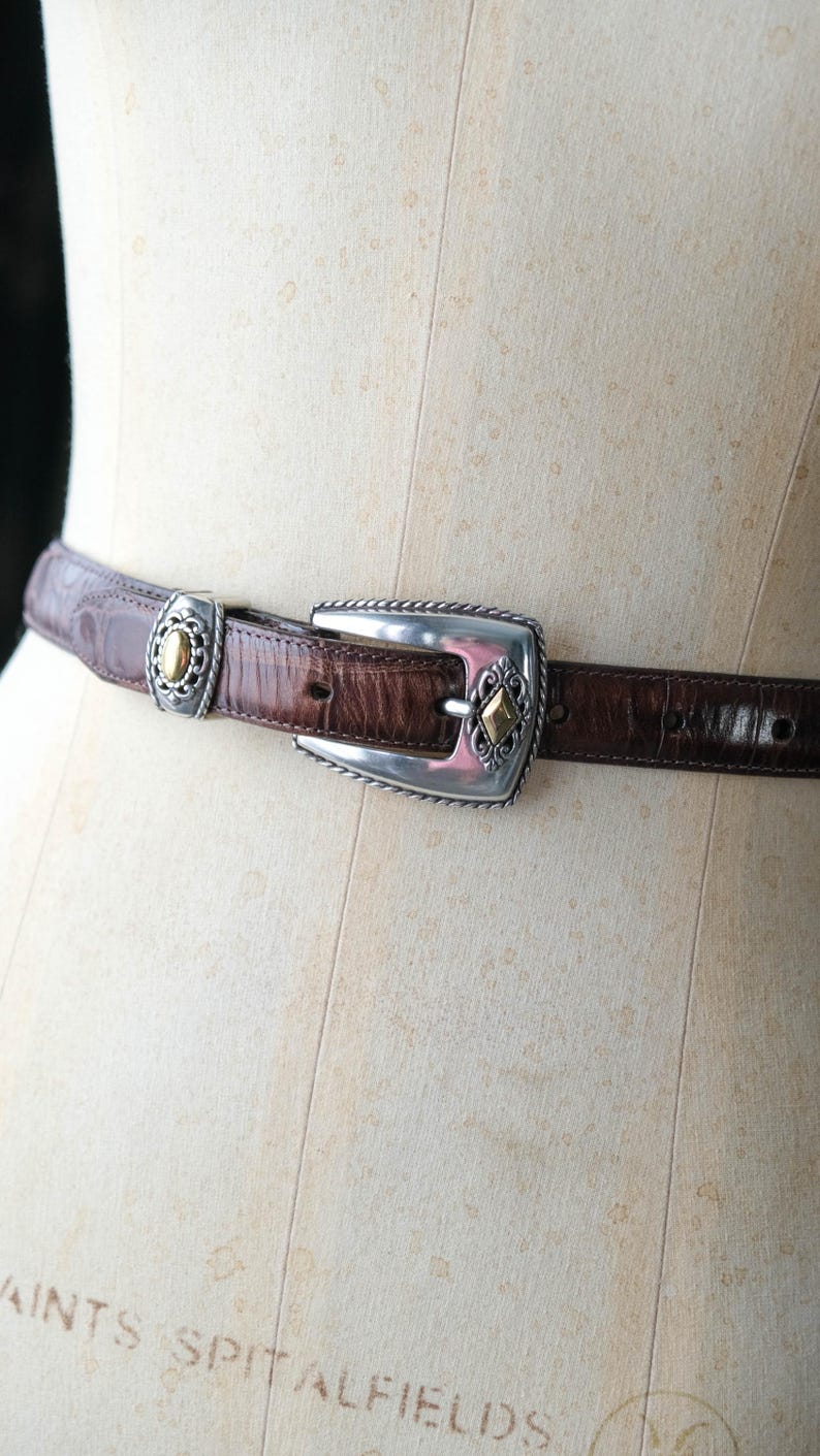 K&ouml;nnte beinhalten: Ein brauner Lederg&uuml;rtel mit silberner Schnalle und Akzenten. Der G&uuml;rtel hat ein strukturiertes, krokodilartiges Muster. Die Schnalle hat einen silbernen Rahmen mit goldfarbenen Details. Der G&uuml;rtel wird auf einer Schaufensterpuppe pr&auml;sentiert.