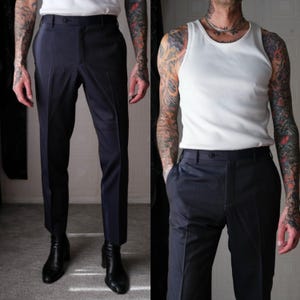 May include: A pair of dark blue tailored pants with a slim fit. The pants have a button fly and belt loops. The pants are shown on a person wearing a white tank top.