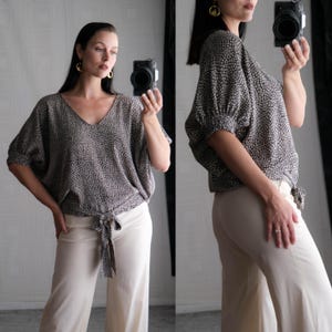 May include: A two-piece outfit featuring a patterned top and cream-colored pants. The top has a V-neck, short sleeves, and a tie at the waist. The pants are wide-legged. The top has a pattern of small, dark dots on a lighter background.