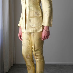 May include: A golden yellow suit with a floral pattern. The suit includes a double-breasted blazer with pockets and matching pants. The outfit is made of a shiny fabric. The person is wearing pink sandals.