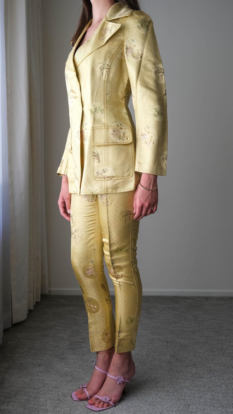 May include: A golden yellow suit with a floral pattern. The suit includes a double-breasted blazer with pockets and matching pants. The outfit is made of a shiny fabric. The person is wearing pink sandals.