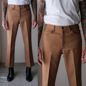 May include: Tan-colored pants with a flared leg design. The pants have a button closure and pockets. The image shows the pants from multiple angles, highlighting the fit and style. The pants are paired with black boots.