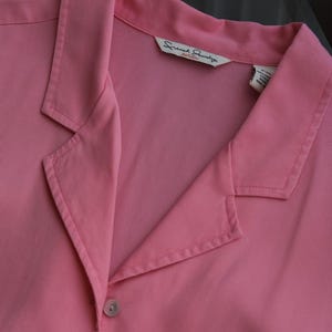 May include: A close-up of a pink collared shirt with two visible buttons. The shirt has a label that reads "French Laundry petite". The shirt is made of a smooth fabric.