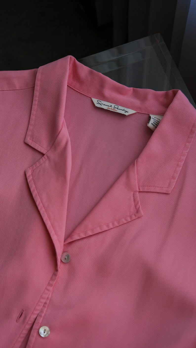 May include: A close-up of a pink collared shirt with two visible buttons. The shirt has a label that reads "French Laundry petite". The shirt is made of a smooth fabric.