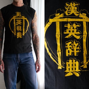 May include: Black sleeveless shirt with a gold graphic of a circle with bamboo stalks and Japanese characters that read "真拳無" (Shin Ken Mu).