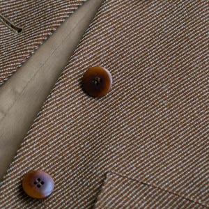 May include: Close-up of a brown tweed blazer with three brown buttons. The blazer has a subtle white stripe pattern and a tan lining visible in the open lapel.