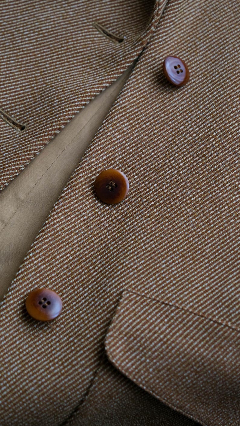 May include: Close-up of a brown tweed blazer with three brown buttons. The blazer has a subtle white stripe pattern and a tan lining visible in the open lapel.