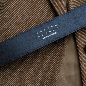 May include: A close-up of a black leather belt with the text "JOSEPH ABBOUD MADE IN ITALY" in white. The belt is positioned over a brown and tan tweed blazer.