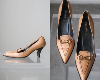 Puede incluir: Zapatos de tacón de cuero color canela con punta afilada y tacón negro de altura media. Los zapatos presentan un detalle de herraje metálico dorado en el empeine y un forro interior negro con el nombre Prada impreso en blanco.
