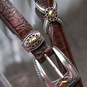 K&ouml;nnte beinhalten: Zwei braune Lederg&uuml;rtel mit silbernen und goldfarbenen Metallschnallen und -akzenten. Die G&uuml;rtel haben ein strukturiertes, krokodilartiges Muster. Eine Schnalle ist ein klassisches Design, die andere hat ein dekoratives Blumenmuster.