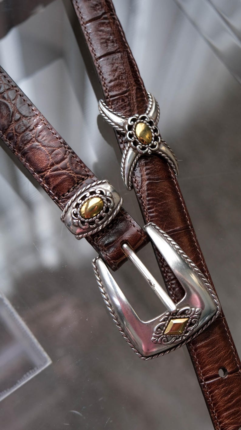 K&ouml;nnte beinhalten: Zwei braune Lederg&uuml;rtel mit silbernen und goldfarbenen Metallschnallen und -akzenten. Die G&uuml;rtel haben ein strukturiertes, krokodilartiges Muster. Eine Schnalle ist ein klassisches Design, die andere hat ein dekoratives Blumenmuster.