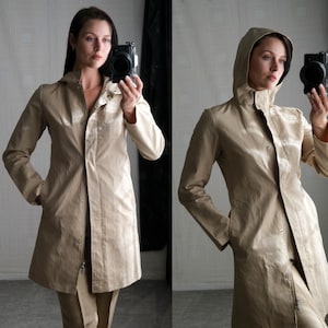 May include: A beige trench coat with a hood and a zipper closure. The coat has long sleeves, pockets, and a tailored fit. The model is wearing the coat and pants. The coat is made of a shiny material.