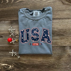 Puede incluir: Una camiseta azul Comfort Colors con la palabra "USA" en letras azul marino con contorno rojo, rellenas de estrellas blancas. El número "1776" está debajo. La camiseta está doblada sobre una superficie de madera.